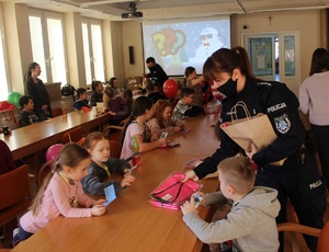 Zdjęcie przedstawia dzieci siedzące przy stole i policjantkę wręczającą elementy odblaskowe.